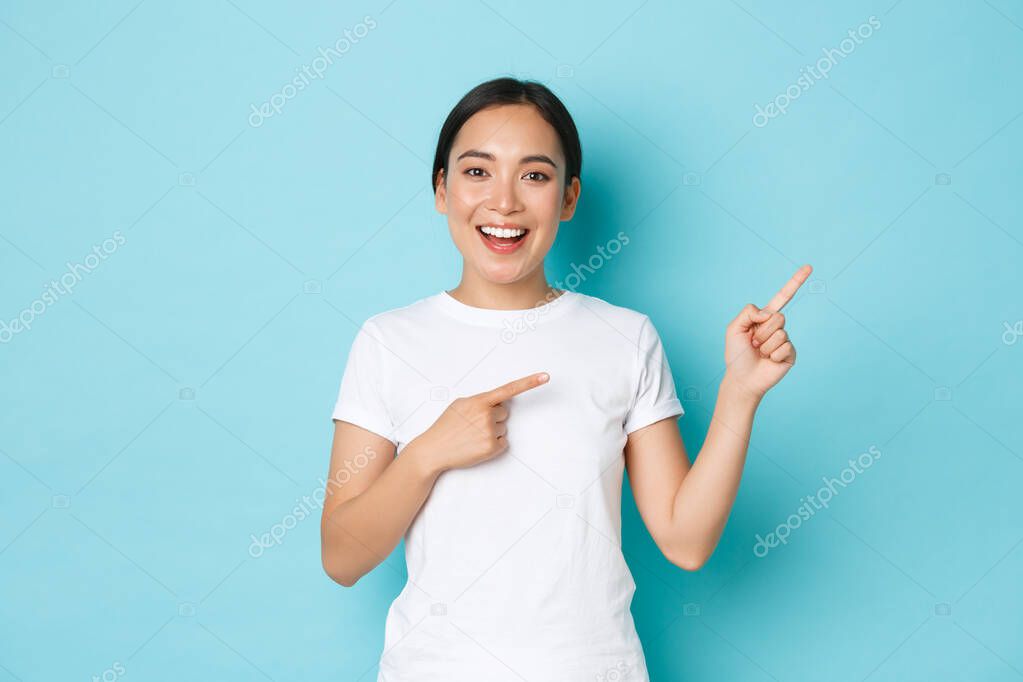 Beautiful happy asian girl in white t-shirt helping with choice, showing way or demonstrating banner. Korean woman looking enthusiastic while pointing upper right corner over blue background