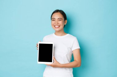 Gururlu ve mutlu gülümseyen Asyalı kız portresi, sevinçle sırıtan, dijital tablet ekranını gösterirken mutlu görünen, iş projesini veya reklamını gösteren, açık mavi arka plan