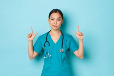 Covid-19, sosyal uzaklık ve koronavirüs salgını konsepti. Şüpheci ve şüpheci genç Asyalı kadın doktor, profesyonel doktor ya da hemşire parmaklarını havaya kaldırarak açık mavi arka plan