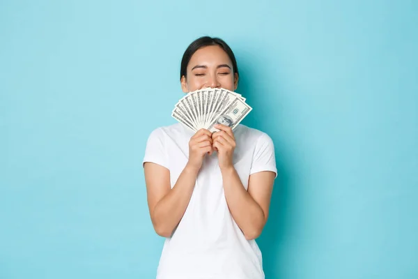 Concepto de compras, dinero y finanzas. Joven exitosa y afortunada chica asiática alegre oliendo dólares y sonriendo satisfecha, disfrutando de ser rica, sosteniendo efectivo sobre fondo azul claro — Foto de Stock