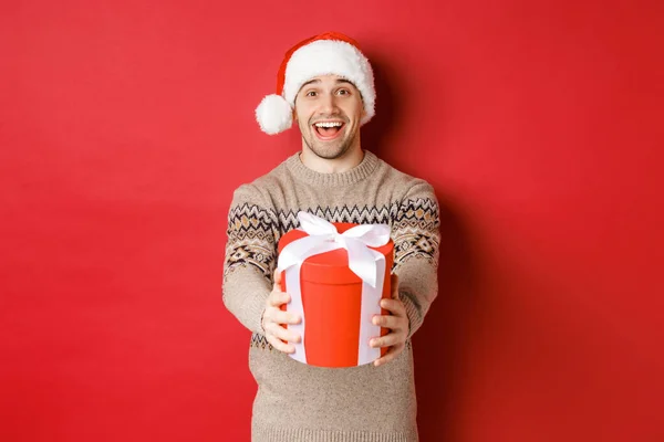 Bild eines glücklichen Mannes, der frohe Weihnachten sagt und Ihnen ein Geschenk in Schachtel schenkt, fröhlich lächelt, in Weihnachtsmütze und Weihnachtspullover die Winterferien feiert, vor rotem Hintergrund steht — Stockfoto