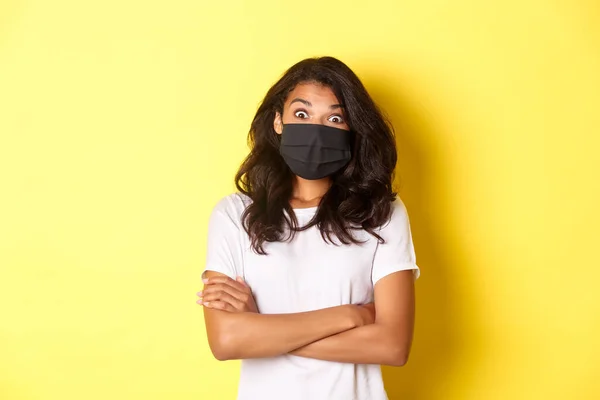 Il concetto di coronavirus, pandemia e stile di vita. Immagine della ragazza afro-americana sorpresa in maschera, guardando stupito qualcosa di fresco, in piedi su sfondo giallo — Foto Stock