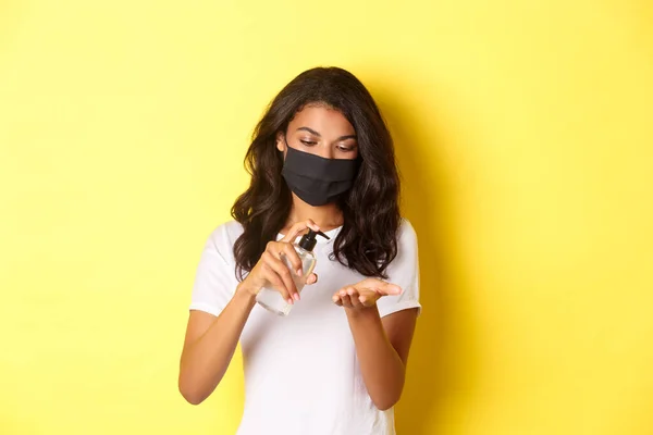 Concepto de covidio-19, distanciamiento social y estilo de vida. Imagen de chica afroamericana en máscara facial usando desinfectante de manos, de pie sobre fondo amarillo — Foto de Stock