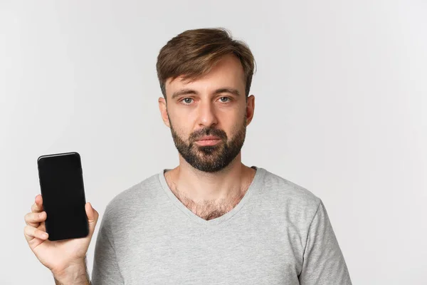 Крупный план уставшего белого парня в серой футболке, выглядящего уставшим и показывающего экран смартфона, стоящего на белом фоне — стоковое фото