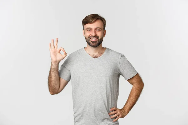 Портрет красивого улыбающегося мужчины в непринужденной серой футболке, показывающий знак одобрения, согласен с вами, восхваляя хороший выбор, стоя на белом фоне — стоковое фото