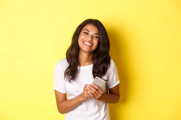 Retrato de la hermosa mujer afroamericana, sonriendo feliz y sosteniendo el teléfono inteligente, de pie sobre el fondo amarillo — Foto de Stock