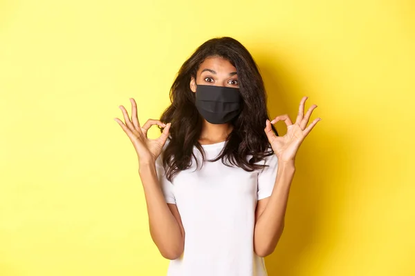 Concepto de coronavirus, pandemia y estilo de vida. Retrato de niña afroamericana complacida en máscara facial negra, mostrando signos de aprobación, como algo bueno, fondo amarillo — Foto de Stock