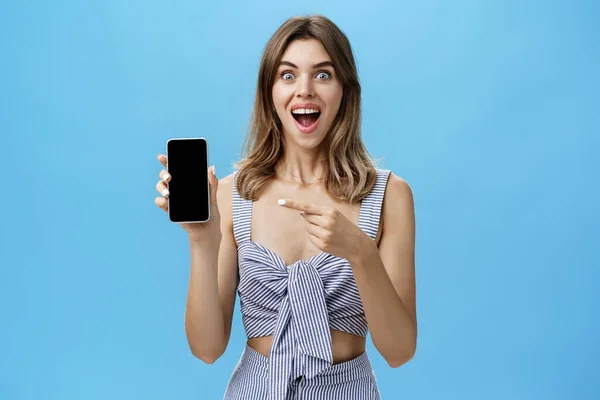 Spännande glad kvinna med gappade tänder äntligen köpt helt ny smartphone innehav enhet i handen pekar på mobiltelefon skärmen visar cool app ler brett från glädje mot blå vägg — Stockfoto