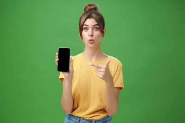 Meraklı kadın portresi bekleyemez. Yeni akıllı telefonu kullan ve cep telefonu ekranını işaret et. Dudakları heyecan ve ilgi ile kıvır ve yeşil duvardaki yeni cihaz modelini tanıt.