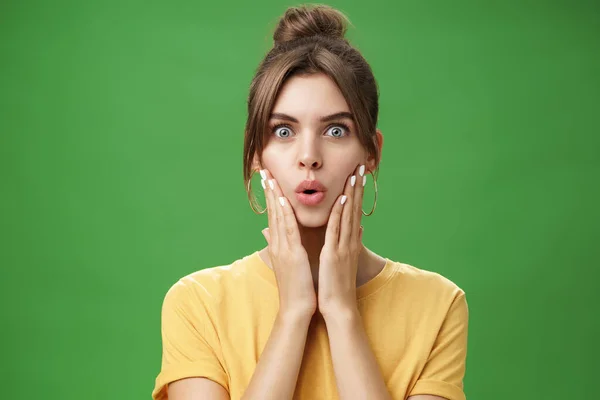 Retrato de mujer femenina linda impresionada y conmocionada en amarillo camiseta plegable labios de la emoción y el interés tocando mejillas sorprendida reaccionando al rumor increíble posando sobre fondo verde — Foto de Stock