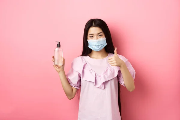 Sağlıklı insanlar ve covid-19 salgını. Tıbbi maskeli neşeli Asyalı kadın el dezenfektanı, baş parmak gösterme, antiseptik şişeyi övme, beyaz arka plan — Stok fotoğraf