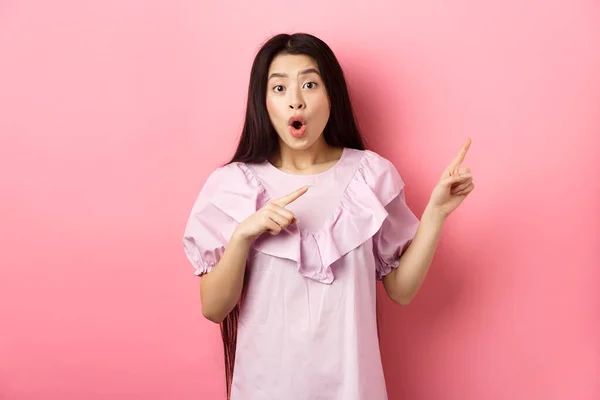 Emocionado mujer asiática mirada asombrada, señalando a la derecha y jadeando fascinado, viendo la publicidad, de pie sobre fondo rosa — Foto de Stock
