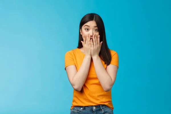 Shocked speechless stunned asian girl stare camera cover mouth palms, raise eyebrows gasping astonished, feel pitty sorry hearing bad news, stand surprised blue background wear yellow t-shirt — Stock Photo, Image