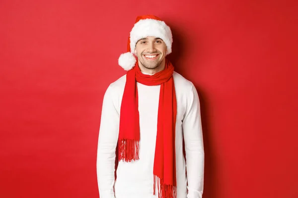 Šťastný běloch slaví Vánoce a usmívá se, nosí Santa klobouk, šálu a bílý svetr, užívá si novoroční prázdniny, stojí nad červeným pozadím — Stock fotografie