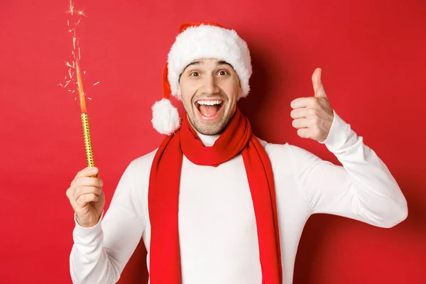 Έννοια των Χριστουγέννων, χειμερινές διακοπές και γιορτή. Όμορφος άνθρωπος γιορτάζει το νέο έτος και τη διασκέδαση, κρατώντας sparkler και δείχνοντας αντίχειρα-up, στέκεται πάνω από το κόκκινο φόντο — Φωτογραφία Αρχείου