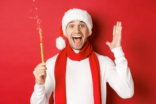 Concept of christmas, winter holidays and celebration. Handsome man celebrating new year and having fun, holding sparkler and smiling, wearing santa hat, standing over red background — Stock Photo, Image