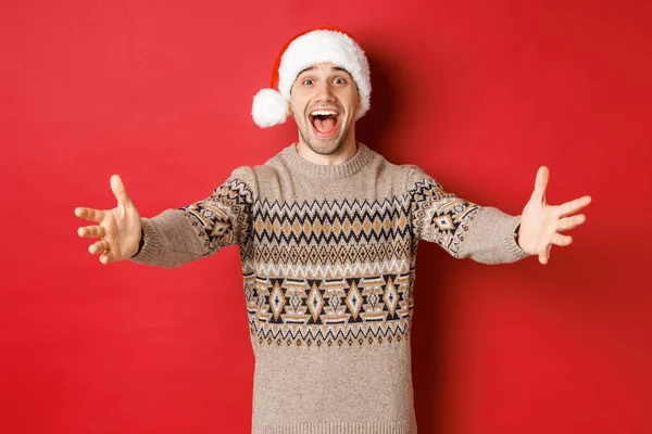 Mutlu, çekici bir adam portresi Noel süveteri ve Noel Baba şapkası giyen, hediye için ellerini uzatan, bir şey alıp gülümseyen, kırmızı arka planda duran — Stok fotoğraf