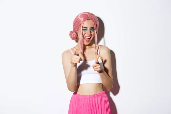 Menina atraente atrevida em traje de Halloween e peruca rosa, apontando para a câmera e sorrindo, parabenizando ou elogiando alguém, de pé sobre fundo branco — Fotografia de Stock
