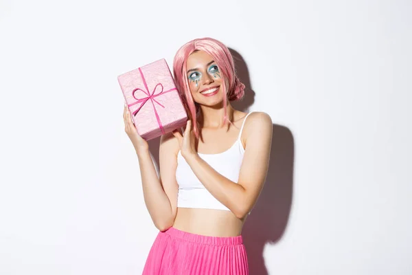 Bild av vacker flicka i rosa peruk skakar låda med födelsedagspresent, undrar vad inuti inslagen låda, står över vit bakgrund — Stockfoto