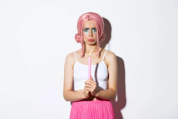 Wizerunek ładnej dziewczyny w różowej peruce dąsającej się rozczarowany po niepowodzeniu, trzymając świecę za magiczne zaklęcie i narzekając, stojąc na białym tle — Zdjęcie stockowe