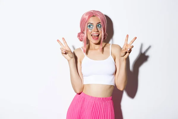 Porträtt av snygg attraktiv flicka i rosa peruk och halloween kostym, visar fred tecken och leende glad, står över vit bakgrund — Stockfoto