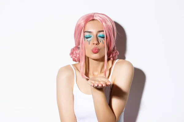 Close-up of lovely girl in halloween costume with pink wig and bright makeup, close eyes and blowing air kiss at camera, standing over white background — Stock Photo, Image