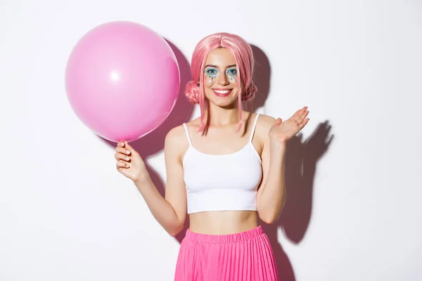 Bild av glad ung flicka med rosa peruk och ljus makeup, håller ballong och viftande hand för att säga hej, hälsning någon på fest, firar semester, vit bakgrund — Stockfoto