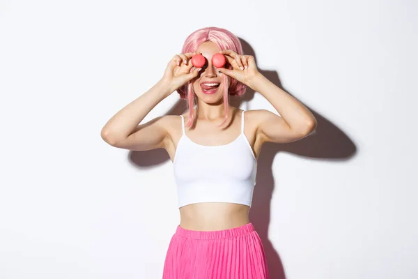 Портрет привабливої дівчини в рожевій вечірці, що тримає макаруни над очима і показує язик з спокусою, стоїть на білому тлі — стокове фото