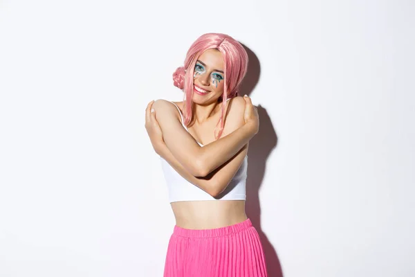 Portrait of beautiful and tender young woman celebrating halloween in glamorous outfit with pink wig, embracing her body and smiling at camera, standing over white background — Stock Photo, Image