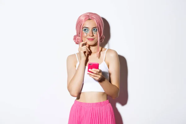 Retrato de niña reflexiva tiene una idea interesante, mirando hacia otro lado y pensando mientras sostiene el teléfono inteligente, usando peluca rosa para la fiesta, celebrando las vacaciones —  Fotos de Stock