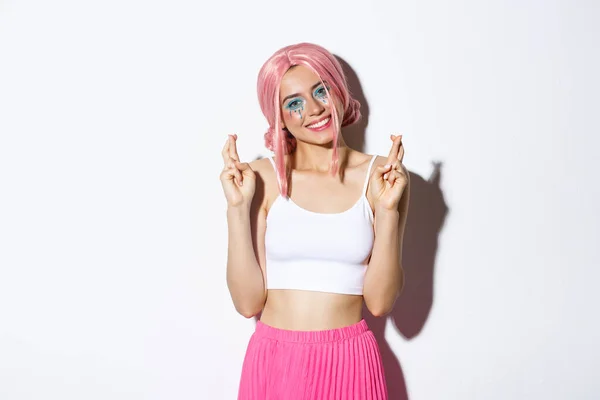 Retrato de mulher sorridente linda em traje de Halloween, peruca rosa e maquiagem brilhante, olhando esperançoso para a câmera e fazendo desejo com os dedos cruzados — Fotografia de Stock