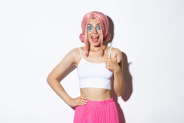Retrato de modelo femenino atractivo entusiasta con pelo rosa y maquillaje de fiesta, mostrando pulgares hacia arriba en la aprobación, sonriendo sorprendido, de pie sobre fondo blanco en traje para halloween —  Fotos de Stock
