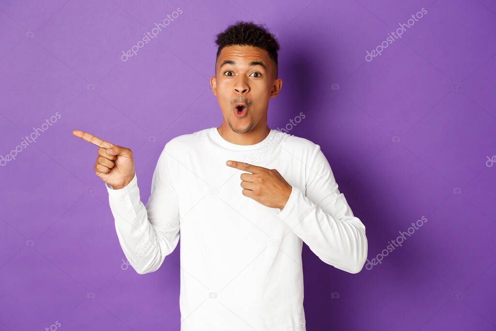 Portrait of amazed african-american man in white sweatshirt, looking impressed and showing advertisement, pointing fingers left, standing over purple background.