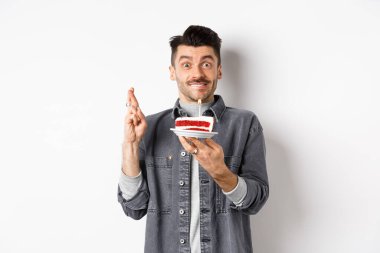 Doğum günü. Mutlu adam dilek diliyor parmakları çapraz, doğum günü pastasını mumla tutuyor, beyaz arka planda duruyor.