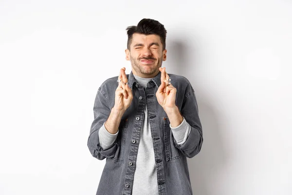 Jeune homme plein d'espoir debout nerveux avec les yeux fermés et les doigts croisés, faisant vœu, suppliant Dieu de réaliser son rêve, attendant des nouvelles, debout sur fond blanc — Photo