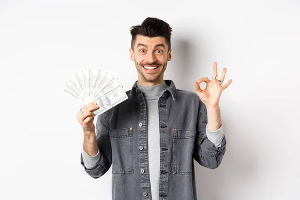Imagen del hombre emocionado sosteniendo billetes de dólar y mostrar signo bien con la cara feliz, haciendo dinero, de pie sobre fondo blanco — Foto de Stock