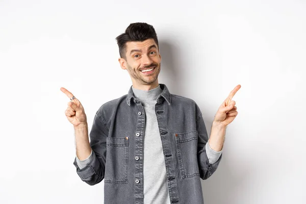 Jonge knappe man met snor wijzende vingers zijwaarts, tonen twee logo 's, lachen blij op de camera, staan in casual kleding op witte achtergrond — Stockfoto