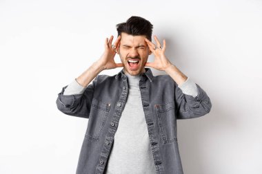 Sinirli genç adam bağırıyor ve kafaya dokunuyor, ayakta duruyor gürültüden ya da baş ağrısından rahatsız oluyor, beyaz arka planda duruyor.
