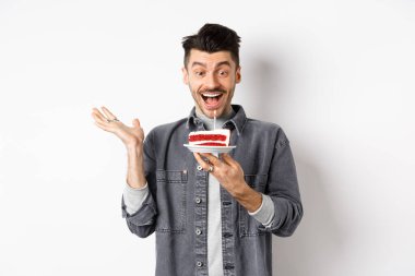 Doğum gününü kutlayan heyecanlı adam, mumla doğum günü pastasına mutlu bakıyor, dilek diliyor, beyaz arka planda mutlu duruyor.