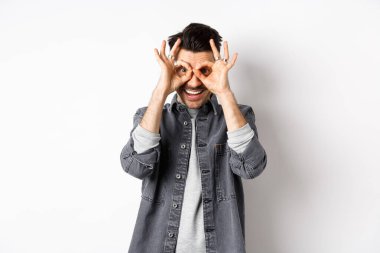 Gülümseyen komik adam, el dürbününden güzel bir teklif görüyor, mutlu ve heyecanlı görünüyor, reklamlara bakıyor, beyaz arka planda duruyor.