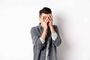 Korkmuş genç adam gergin yüzüyle parmaklarının arasından bakıyor, korkunç bir şeye bakıyor, beyaz arka planda duruyor.