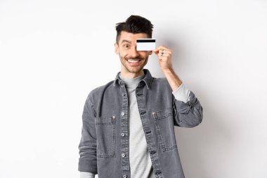 Bıyıklı mutlu yakışıklı adam gözlerinde plastik kredi kartı, kameraya heyecanlı bir şekilde gülümsüyor, beyaz arka planda duruyor.