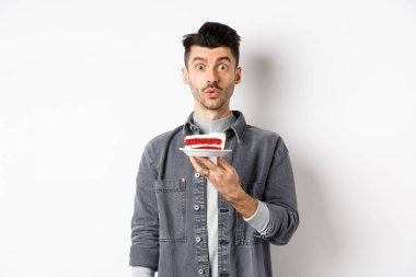 Yakışıklı doğum günü çocuğu pastaya mum üflüyor ve dilek tutuyor, kameraya bakıp, beyaz arka planda duruyor.