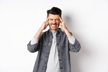 Baş ağrısı çeken, baş ağrısından dişlerini sıkan, baş şakaklarına dokunan, migren ağrısı çeken, beyaz arka planda duran, ağrı kesicilere ihtiyacı olan bir adam.