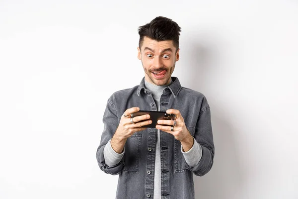 Homme excité jouant à un jeu vidéo sur téléphone portable avec le visage concentré, appuyez sur l'écran du smartphone, tenant le téléphone portable horizontalement, fond blanc — Photo