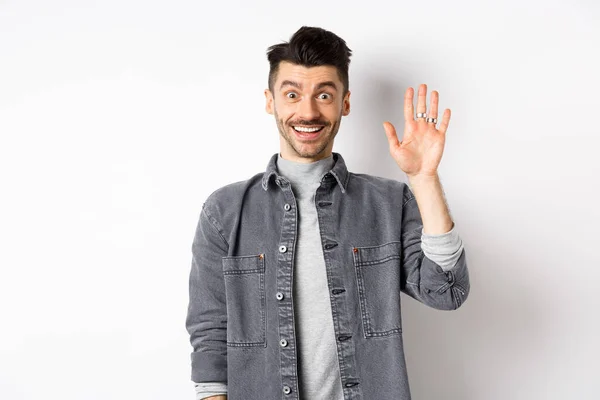 Vriendelijke stijlvolle man zegt hallo en aarzelende hand, glimlachend vrolijk, begroeten u met hi gebaar, staan op witte achtergrond — Stockfoto