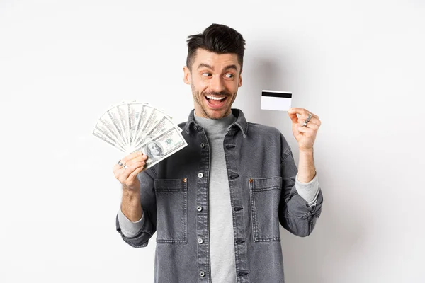 Tipo excitado sosteniendo tarjetas de crédito de plástico y billetes de dólar, de pie divertido sobre fondo blanco — Foto de Stock