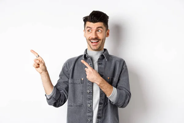 잘생긴 미소짓는 남자가 광고를 확인하고 흥분 한 얼굴로 왼쪽을 보고 지적 백인 배경 위에 서서 — 스톡 사진