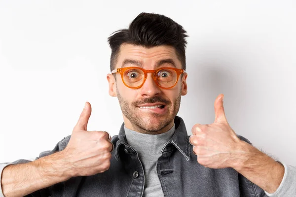 Nervöser junger Mann beißt auf die Lippe und zeigt Daumen hoch, trägt stylische Brille, steht vor weißem Hintergrund — Stockfoto