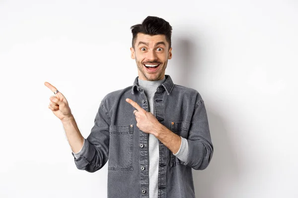 Tipo excitado con bigote señalando los dedos a la izquierda, mostrando publicidad y sonriendo feliz, de pie sobre fondo blanco — Foto de Stock